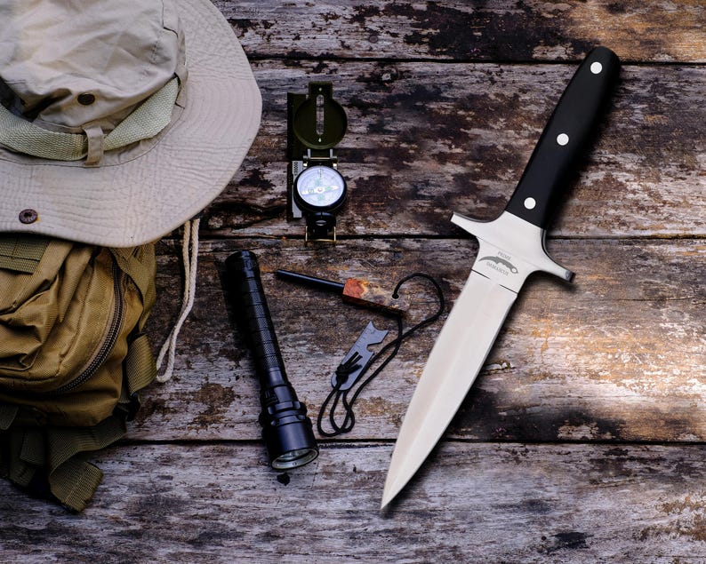 May include: A collection of outdoor survival gear including a knife with a black handle and silver blade, a compass, a torch, a fire starter, a beige hat, and a backpack. The items are arranged on a weathered wooden surface.