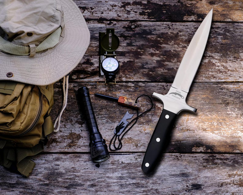 May include: An outdoor adventure kit featuring a survival knife with a black handle, a compass, a flashlight, a fire starter, a tan backpack, and a beige hat. The items are arranged on a weathered wooden surface.
