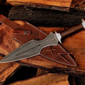May include: A handcrafted dagger with a dark, textured blade and a wooden handle. The dagger rests in a brown leather sheath with decorative stitching. The handle has a silver bolster. The dagger is displayed on a wooden surface.