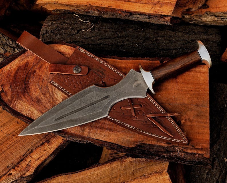 May include: A handcrafted dagger with a dark, textured blade and a wooden handle. The dagger rests in a brown leather sheath with decorative stitching. The handle has a silver bolster. The dagger is displayed on a wooden surface.