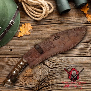 May include: A brown leather sheath covers a large hunting knife with a wooden handle. The knife and sheath rest on a wooden surface, alongside a coil of rope, a green hat, and a few leaves. The sheath has an embossed design.