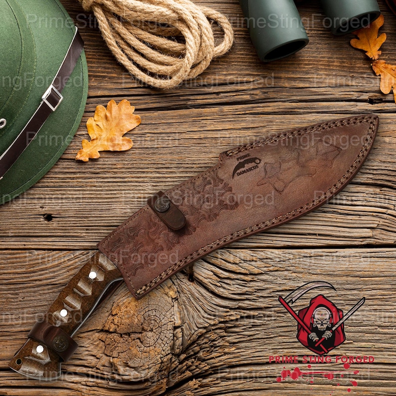 May include: A brown leather sheath covers a large hunting knife with a wooden handle. The knife and sheath rest on a wooden surface, alongside a coil of rope, a green hat, and a few leaves. The sheath has an embossed design.
