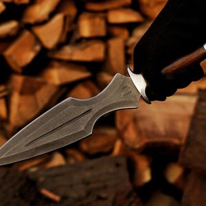May include: A dark grey dagger with a pointed blade and a decorative handle. The handle is made of wood and metal. The dagger is held by a gloved hand against a background of stacked wood.