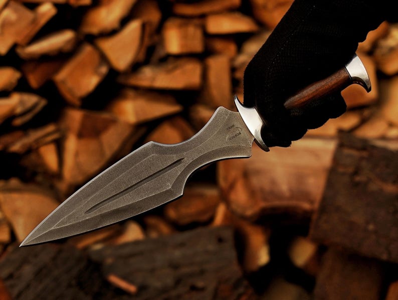 May include: A dark grey dagger with a pointed blade and a decorative handle. The handle is made of wood and metal. The dagger is held by a gloved hand against a background of stacked wood.