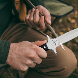 May include: A silver and black hunting knife with a sharp blade is held. The knife has a black handle and a silver blade. The person is sitting outdoors, sharpening a wooden stick with the knife.