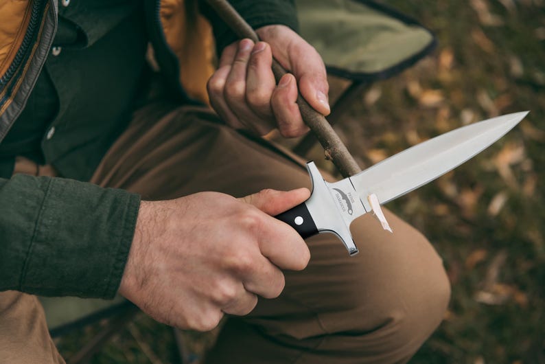 May include: A silver and black hunting knife with a sharp blade is held. The knife has a black handle and a silver blade. The person is sitting outdoors, sharpening a wooden stick with the knife.