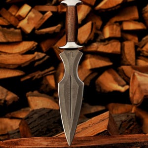 May include: A dagger with a dark, textured blade and a wooden handle. The handle is capped with a silver-coloured metal. The dagger is displayed against a backdrop of stacked firewood, creating a rustic aesthetic.