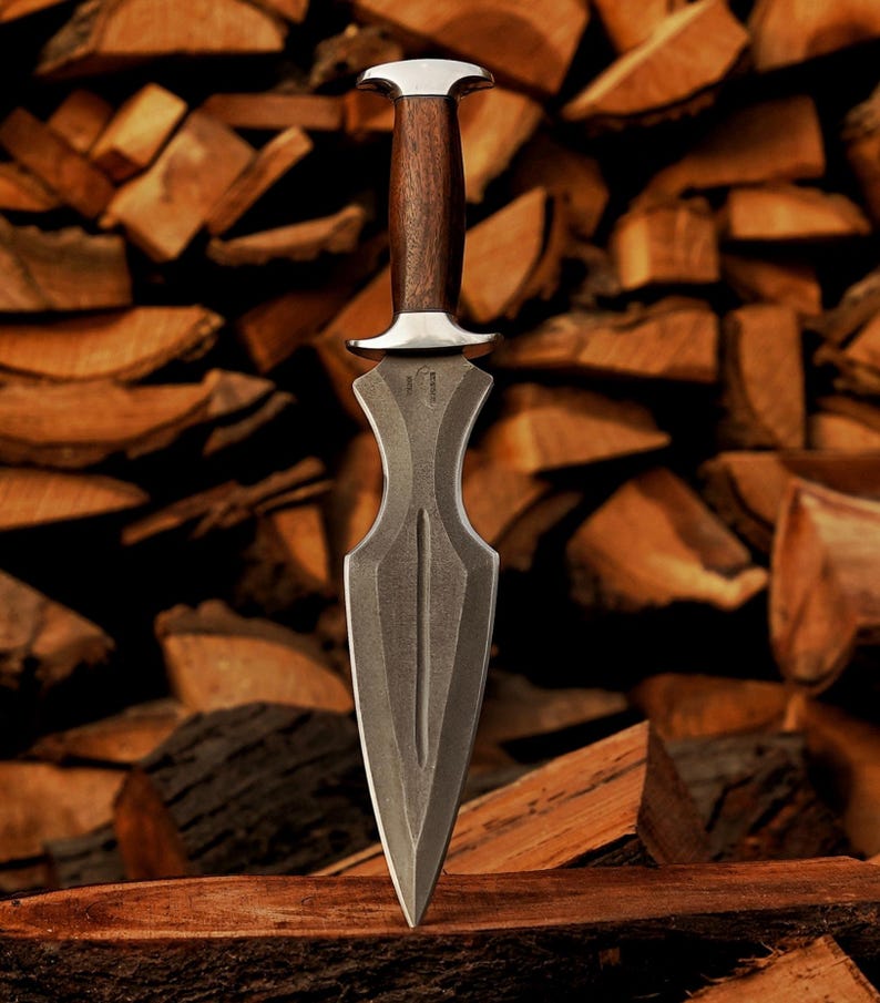 May include: A dagger with a dark, textured blade and a wooden handle. The handle is capped with a silver-coloured metal. The dagger is displayed against a backdrop of stacked firewood, creating a rustic aesthetic.