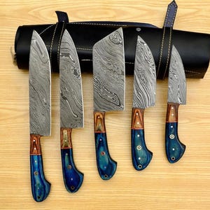 May include: Set of five kitchen knives with blue and brown handles and a leather roll for storage. The knives have a Damascus steel blade pattern.