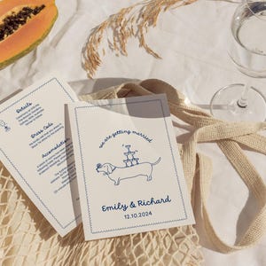 May include: A white wedding invitation with blue text and a cartoon illustration of a dachshund carrying a stack of champagne glasses. The text reads "we are getting married" and "Emily & Richard 12.10.2024".