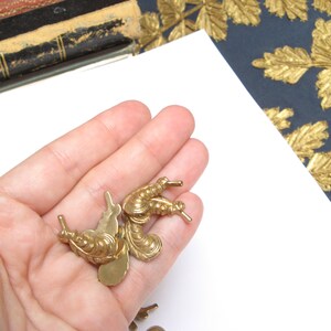 May include: Several small, ornate, gold-colored metal embellishments are held in a hand. The embellishments have a detailed, vintage design with curved lines and small protrusions. The background includes a white surface, a dark blue surface with gold leaf designs, and a glimpse of a book.