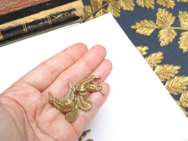 May include: Several small, ornate, gold-colored metal embellishments are held in a hand. The embellishments have a detailed, vintage design with curved lines and small protrusions. The background includes a white surface, a dark blue surface with gold leaf designs, and a glimpse of a book.