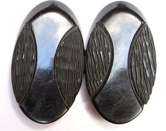 May include: A two-piece oval belt buckle in a glossy black finish. Each oval section features a smooth, reflective central panel flanked by two textured, carved side panels with vertical grooves. The pieces are joined by a central metal hinge.