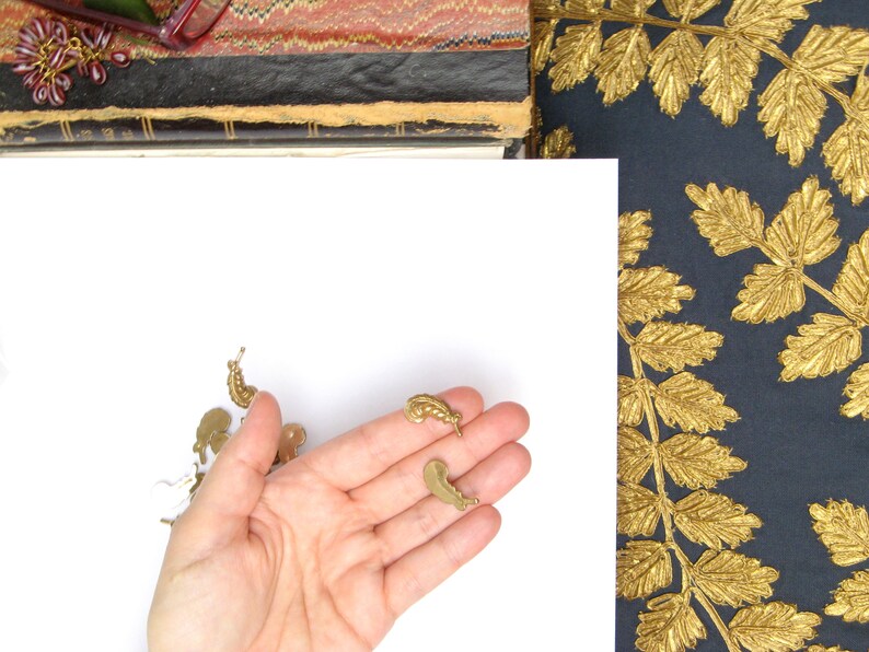 May include: A collection of small, brass-colored charms in various shapes, including snails and other organic forms, are displayed against a white background. The charms are held in a hand. A dark blue fabric with gold leaf designs is visible in the background.
