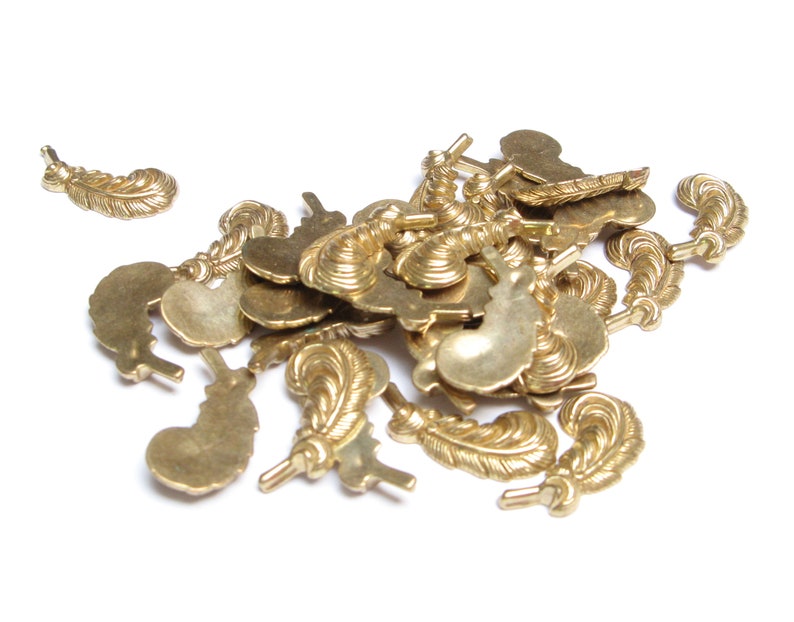 May include: A collection of small, gold-colored feather charms. The charms have detailed feather designs and are scattered on a white surface. These charms are suitable for jewelry making or craft projects.