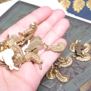 May include: A collection of small, ornate brass feather-shaped embellishments. Some are solid, while others have a cutout design. The feathers are held in a hand, with a vintage book visible in the background. These decorative elements are ideal for crafting and jewelry making.
