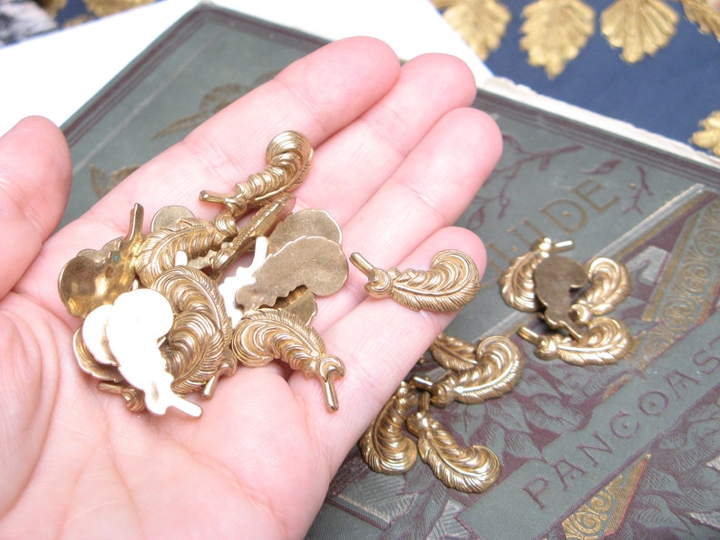 May include: A collection of small, ornate brass feather-shaped embellishments. Some are solid, while others have a cutout design. The feathers are held in a hand, with a vintage book visible in the background. These decorative elements are ideal for crafting and jewelry making.