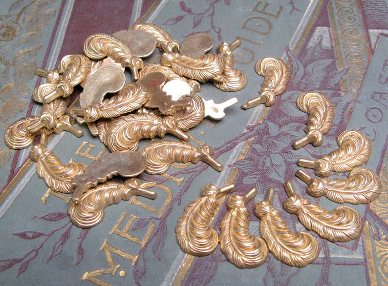 May include: A collection of gold-colored feather-shaped metal embellishments. The feathers have detailed veining and a small post at the base. They are scattered on a background with gold lettering and floral designs.