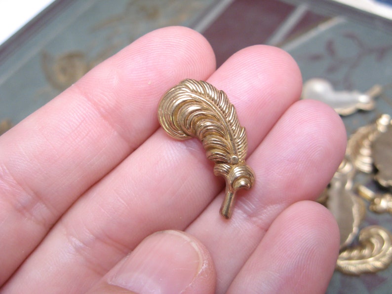 May include: A small, gold-colored feather-shaped embellishment. The feather has detailed veining and a curved top, with a small stem at the base. Other decorative metal pieces are visible in the background.