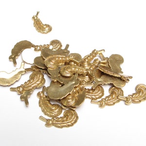 May include: A collection of small, brass-colored feather charms scattered on a white surface. The charms have detailed feather designs and small loops for attaching to jewelry or other crafts. The feathers vary in size and orientation.