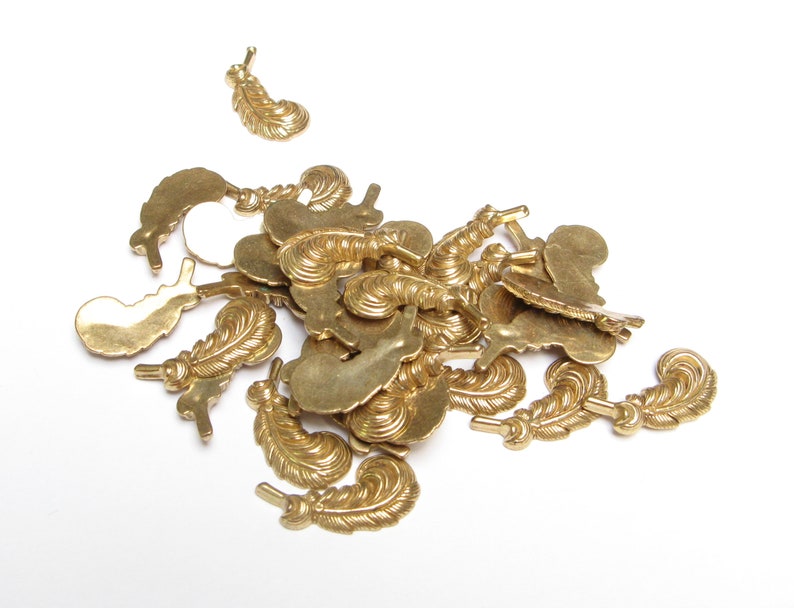 May include: A collection of small, brass-colored feather charms scattered on a white surface. The charms have detailed feather designs and small loops for attaching to jewelry or other crafts. The feathers vary in size and orientation.