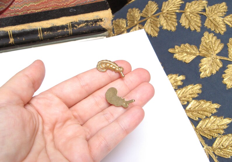 May include: Two small, gold-colored metal charms in the shape of a feather and a snail are held in a hand. The charms are on a white surface, with a dark blue background featuring gold leaf designs.