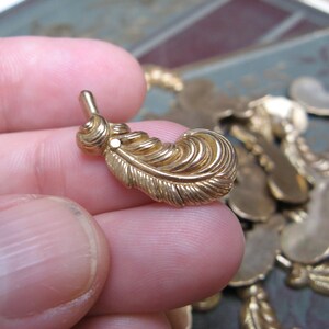 May include: A gold-colored feather charm with intricate detailing. The charm has a small loop at the top and a detailed feather design. Other similar charms are visible in the background. This charm is suitable for jewelry making.