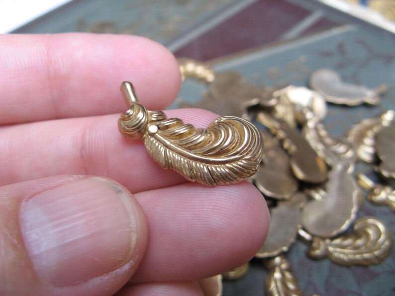 May include: A gold-colored feather charm with intricate detailing. The charm has a small loop at the top and a detailed feather design. Other similar charms are visible in the background. This charm is suitable for jewelry making.