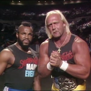 May include: Two men stand side-by-side, both wearing sleeveless shirts. The man on the left has a mohawk and is wearing a black shirt with red and blue text that reads "Saturday Night's Main Event". The man on the right has long blonde hair and is wearing a black tank top with red text that reads "Saturday Night's". He is also wearing a gold championship belt.