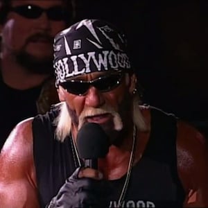 May include: A man wearing a black bandana with the word "HOLLYWOOD" in white letters, black sunglasses, and a black tank top. He is holding a microphone and speaking. Another man wearing sunglasses is in the background.