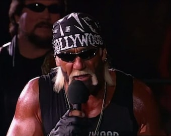 May include: A man wearing a black bandana with the word "HOLLYWOOD" in white letters, black sunglasses, and a black tank top. He is holding a microphone and speaking. Another man wearing sunglasses is in the background.