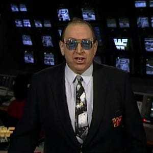 Könnte beinhalten: Ein Mann in einem schwarzen Anzugjackett mit weißem Hemd und einer gemusterten Krawatte. Er trägt eine Sonnenbrille und hat ein Mikrofon am Revers. Der Hintergrund ist eine Wand mit Fernsehbildschirmen, auf denen das WWF-Logo zu sehen ist.