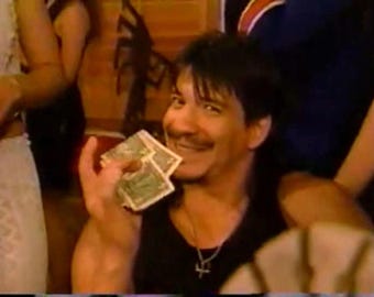 May include: A man with dark hair and a goatee is smiling and holding a stack of US dollar bills. He is wearing a black tank top and a silver necklace with a cross pendant.