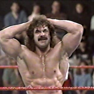 May include: A muscular man with long brown hair and a moustache is standing in a wrestling ring. He is wearing a black wrestling singlet and has his hands on his head. He is looking down at the ground.
