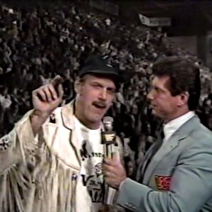 May include: A retro image of two men in a wrestling arena. One man is wearing a white jacket with fringe and a black cap, holding a microphone. The other man is wearing a light blue suit. A crowd fills the background.