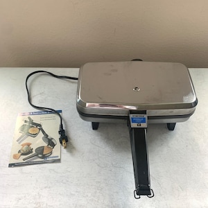 May include: A Vitantonio Pizzelle Chef electric iron with a rectangular stainless steel top, a black handle, and a black power cord. A printed recipe and instruction booklet is included, featuring images of the appliance and pizzelle cookies.
