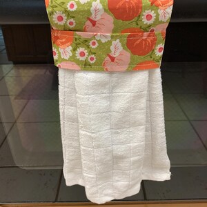 May include: A kitchen towel holder with a white towel hanging from it. The holder features a green fabric with a pattern of orange pumpkins, pink pumpkins, and white flowers. The holder is attached to a black oven handle.