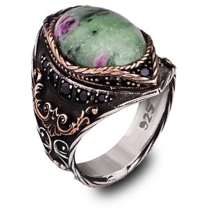 May include: A silver ring with a green and pink gemstone set in a gold-toned bezel. The ring is decorated with black stones and intricate details. The ring is stamped with "925" on the side.
