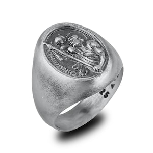 May include: A silver ring with a raised oval design featuring a depiction of Saint Christopher carrying a child. The ring is engraved with the words "St. Christopher" and the number "25".