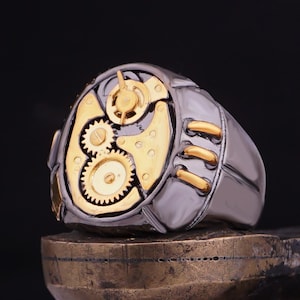 May include: A silver and gold ring with a steampunk design featuring gears and cogs. The ring is set on a brown surface.