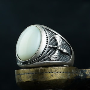 May include: A silver ring with an oval, off-white stone. The ring features a detailed design of a bird with outstretched wings on one side. The ring is set on a gold-colored surface against a dark background.