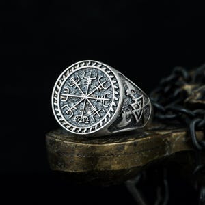 May include: A silver-colored ring with a circular face featuring a detailed Viking compass design. The ring has a textured band and is displayed on a dark surface, showcasing its intricate craftsmanship. The ring is a piece of jewelry.