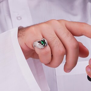 May include: A silver ring featuring a design of two hands cradling a large, oval-cut, emerald-green gemstone. The ring is worn on a finger, with the hand and part of a white shirt visible in the image.