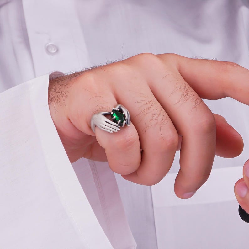 May include: A silver ring featuring a design of two hands cradling a large, oval-cut, emerald-green gemstone. The ring is worn on a finger, with the hand and part of a white shirt visible in the image.