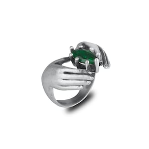 May include: A silver ring featuring two hands cradling a large, oval-cut, emerald-green gemstone. The ring has a detailed, textured finish, and the hands are sculpted with realistic features. The ring is set against a white background.