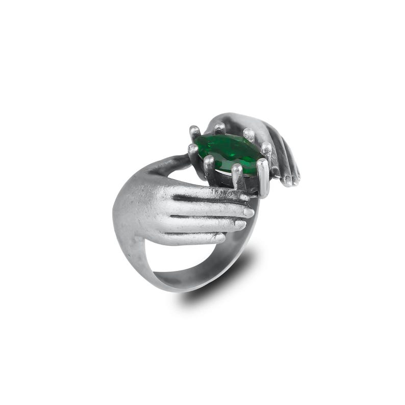 May include: A silver ring featuring two hands cradling a large, oval-cut, emerald-green gemstone. The ring has a detailed, textured finish, and the hands are sculpted with realistic features. The ring is set against a white background.