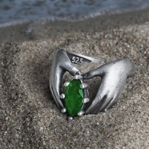 May include: A sterling silver ring featuring a green gemstone held by two sculpted hands. The ring is set against a sandy background with the ocean visible in the distance. The ring is stamped with "925".