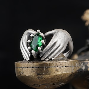 May include: A silver sculpture of two hands cradling a large, faceted emerald-green gemstone. The hands are detailed with textured surfaces, set on a bronze-colored base against a black background. The sculpture is a decorative art piece.