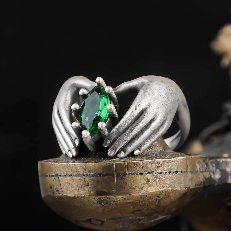 May include: A silver sculpture of two hands cradling a large, faceted emerald-green gemstone. The hands are detailed with textured surfaces, set on a bronze-colored base against a black background. The sculpture is a decorative art piece.