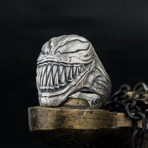 May include: A silver-colored ring featuring a detailed, sculpted design of a monster's face with sharp teeth. The ring is set against a dark background, highlighting the intricate details of the metalwork. The ring is a unique piece of jewelry.